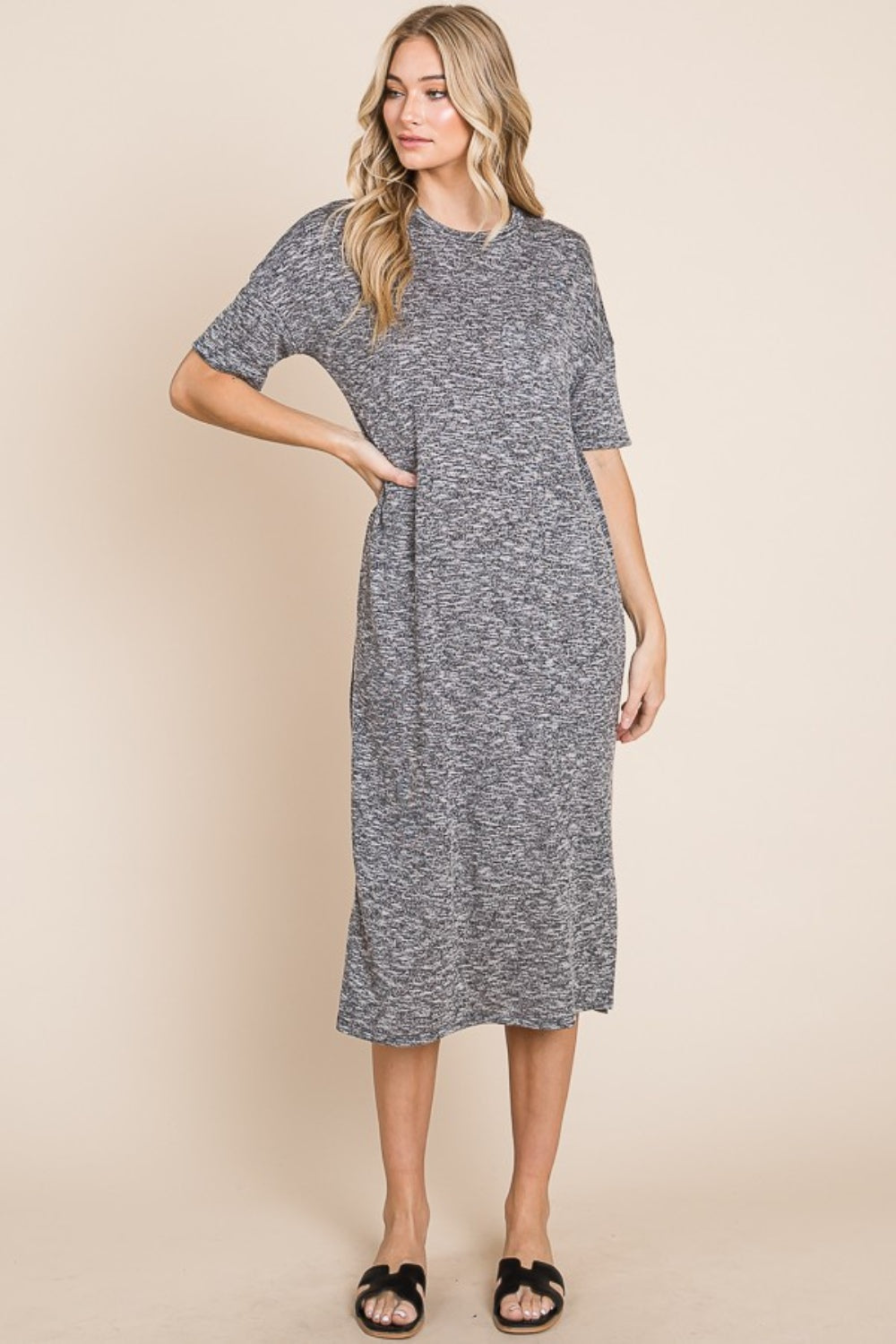 A woman wearing the BOMBOM Slit Round Neck Half Sleeve Midi Dress, a knee-length, grey midi dress featuring short sleeves and a slit detail at the hem, paired with black slip-on sandals, stands in front of a neutral background.