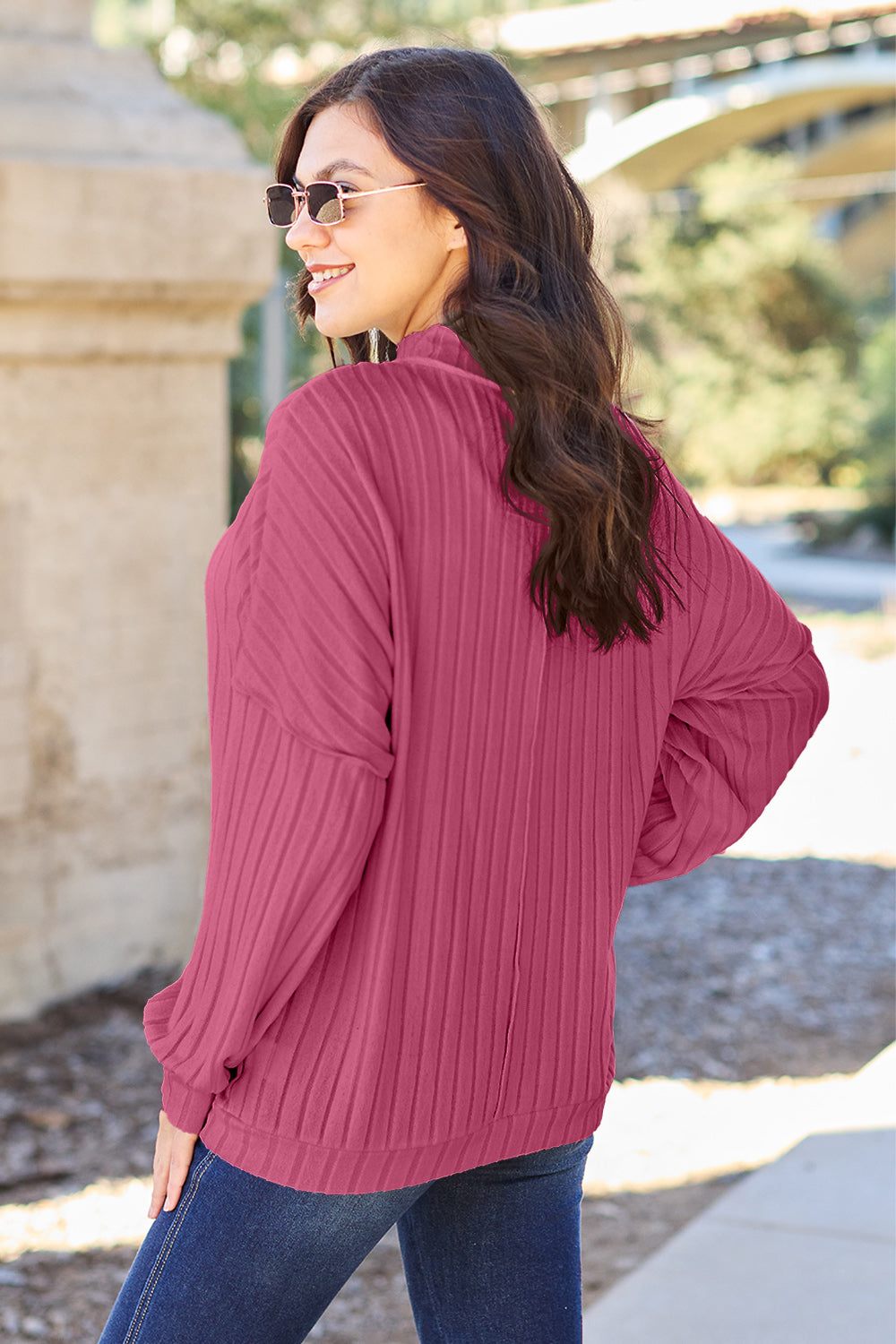 A woman with long hair is wearing a green Basic Bae Full Size Ribbed Exposed Seam Mock Neck Knit Top, blue jeans, and sunglasses. She is standing outdoors and smiling.