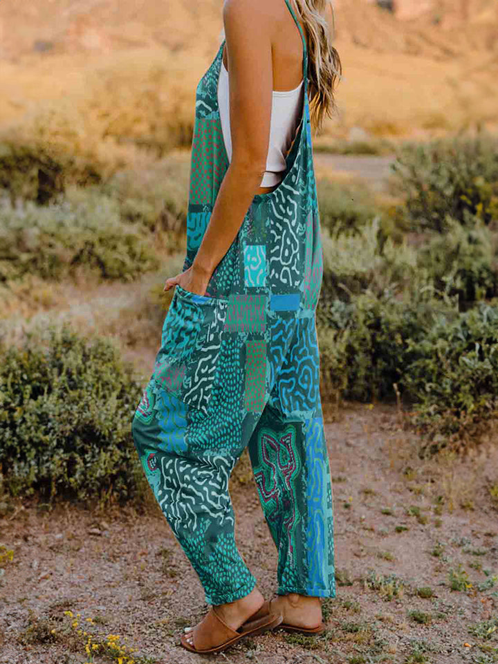 A person standing outdoors in front of rocky terrain, wearing a Full Size Printed V-Neck Sleeveless Jumpsuit and a hat.