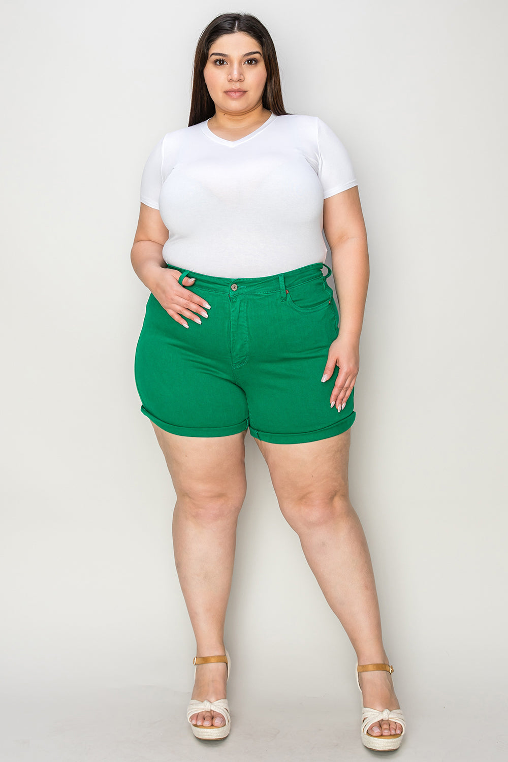 Two women stand indoors wearing white tops and Judy Blue Full Size Tummy Control Garment Dyed Denim Shorts in green. The woman on the left has her hand in her pocket, and both are smiling. There are plants and decorations in the background.