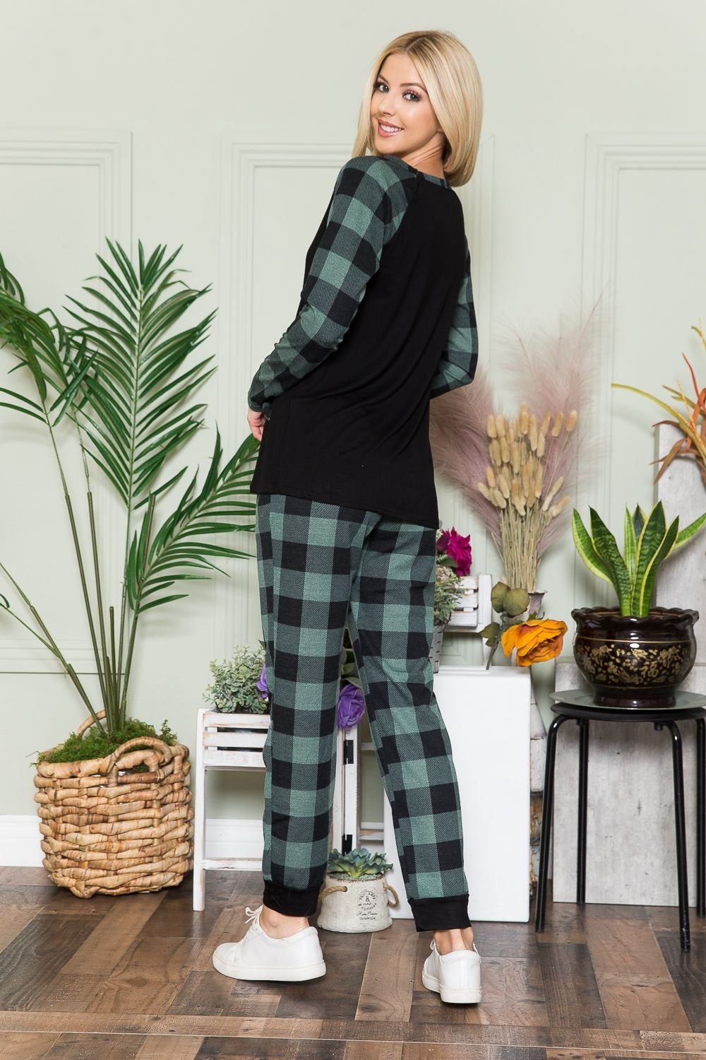 An individual wearing a Celeste Plaid Long Sleeve T-Shirt with Pockets stands indoors on a wooden floor, next to potted plants and a wicker basket.