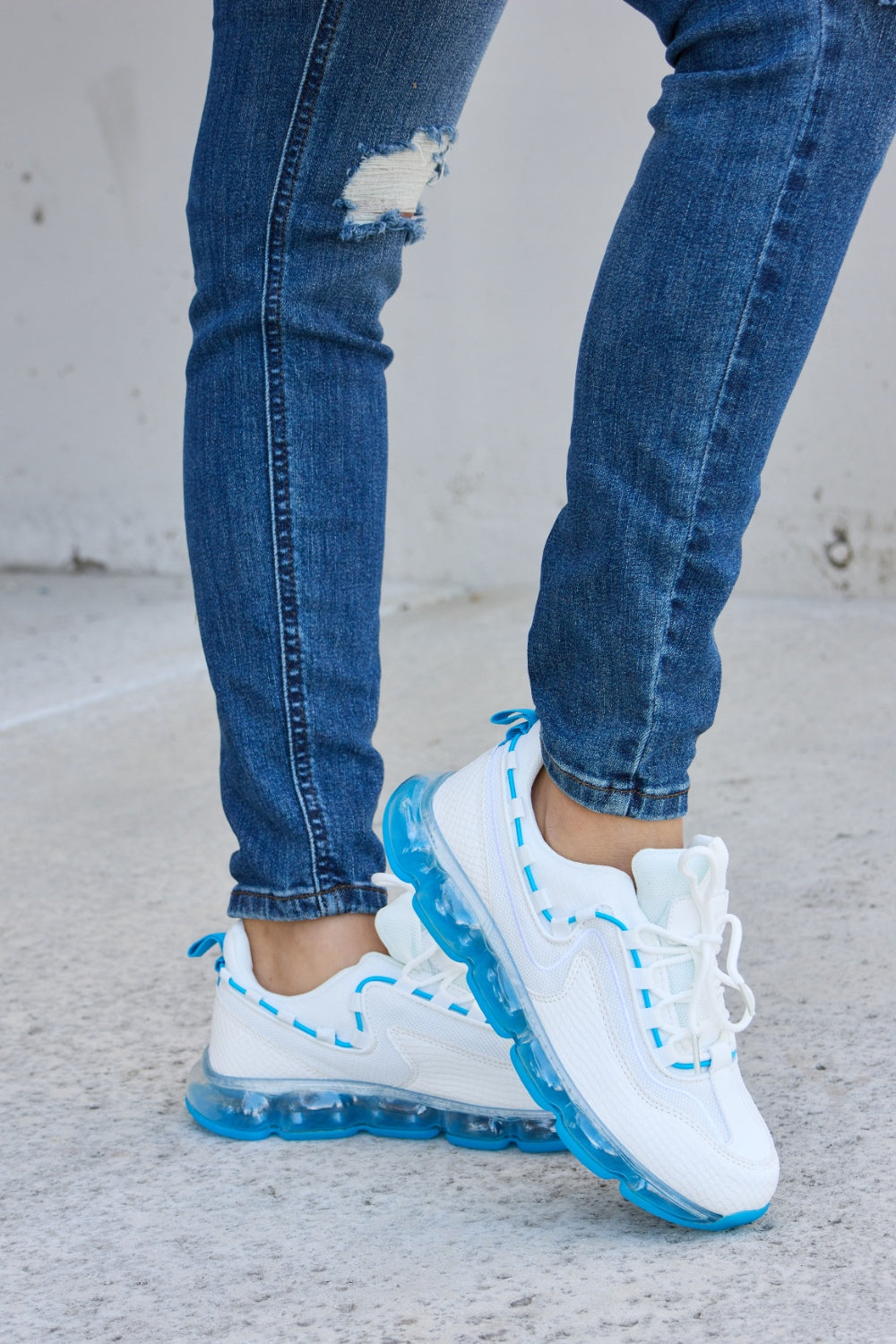 A person wearing ripped jeans and stylish Forever Link Lace-Up Air-Cushioned Athletic Shoes in white with blue accents stands confidently on a concrete surface.