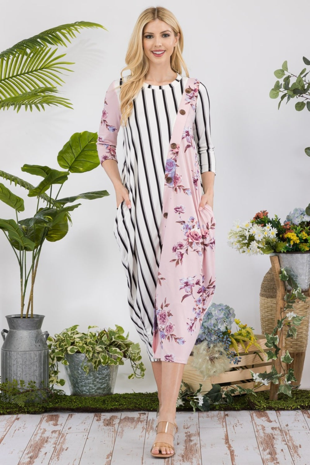 A woman stands amidst potted plants and flowers on a wooden floor, wearing the elegant and chic Celeste Full Size Floral Striped Contrast Midi-Dress with Pockets.