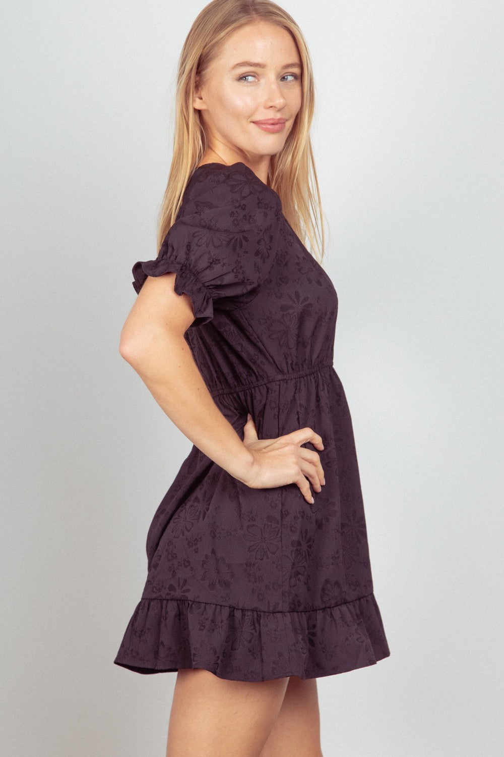 Person in a VERY J Floral Textured Woven Ruffled Mini Dress standing and smiling with one hand on hip.