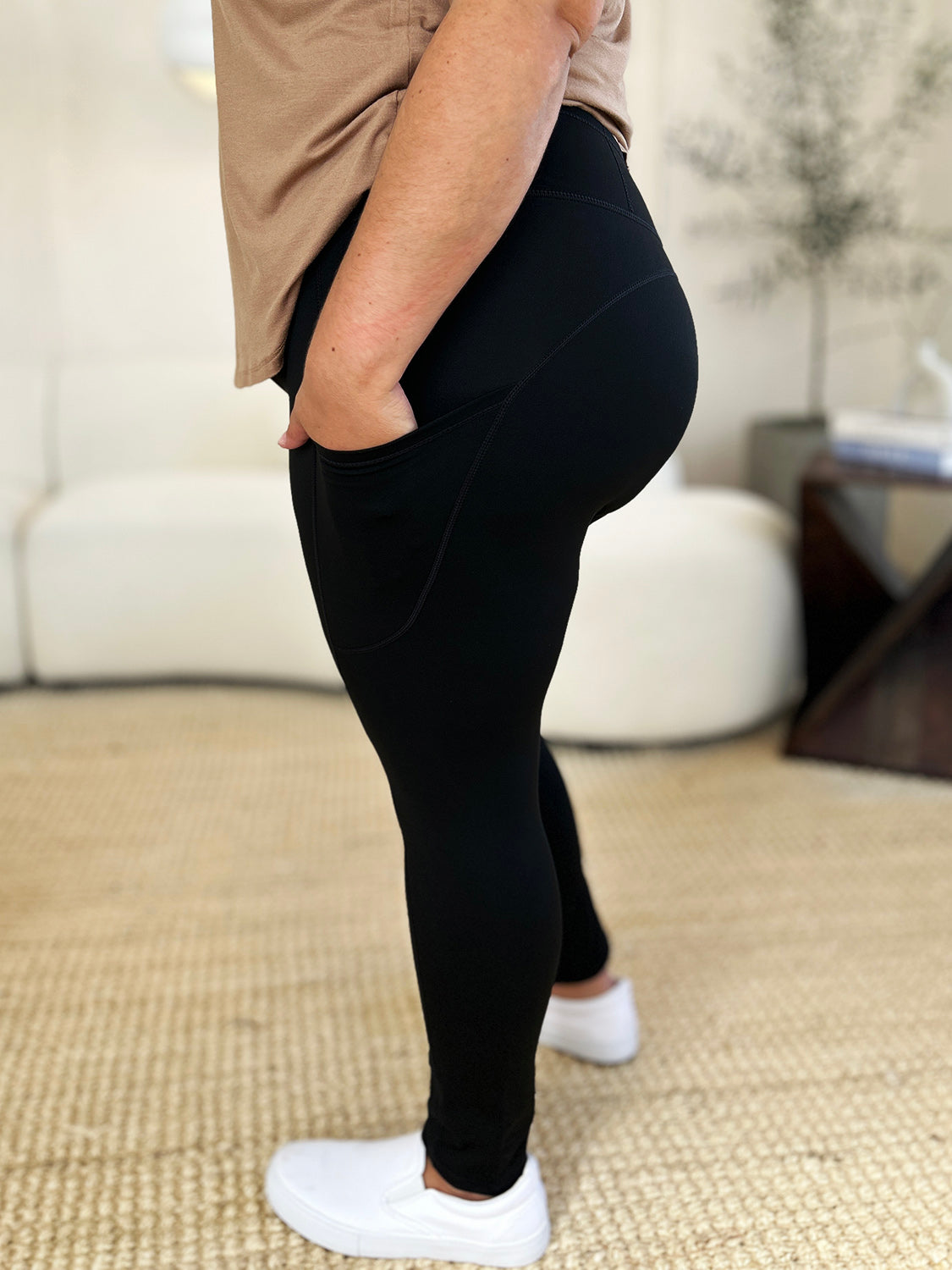 Person wearing Wide Waistband Sports Leggings made of stretchy nylon spandex and white slip-on shoes, standing on a beige carpet with a white couch in the background.