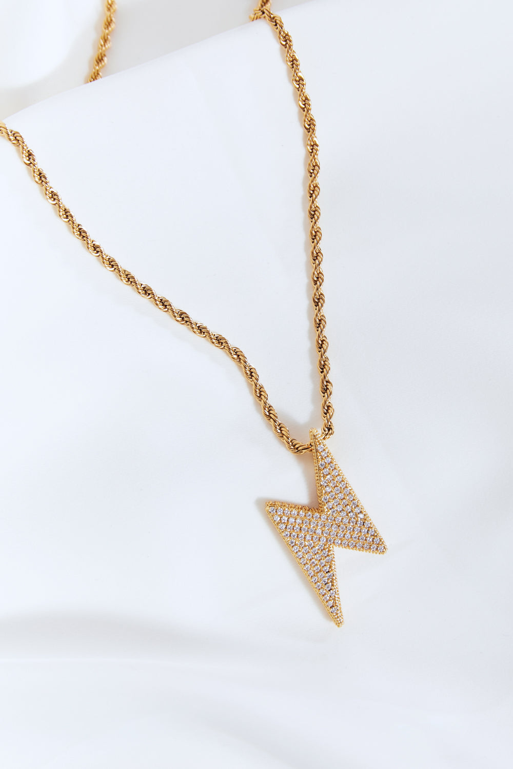 A Brass Lightning Pendant Necklace, featuring a gold twisted chain and a lightning bolt-shaped pendant encrusted with small cubic zirconia stones, displayed on a light blue background.