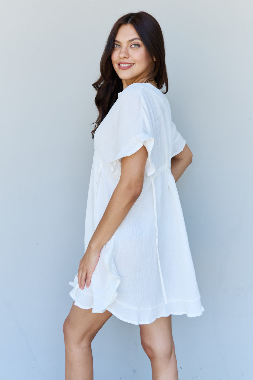 A person with long dark hair smiles while posing in the Ninexis Out Of Time Full Size Ruffle Hem Dress with Drawstring Waistband in White against a plain background.