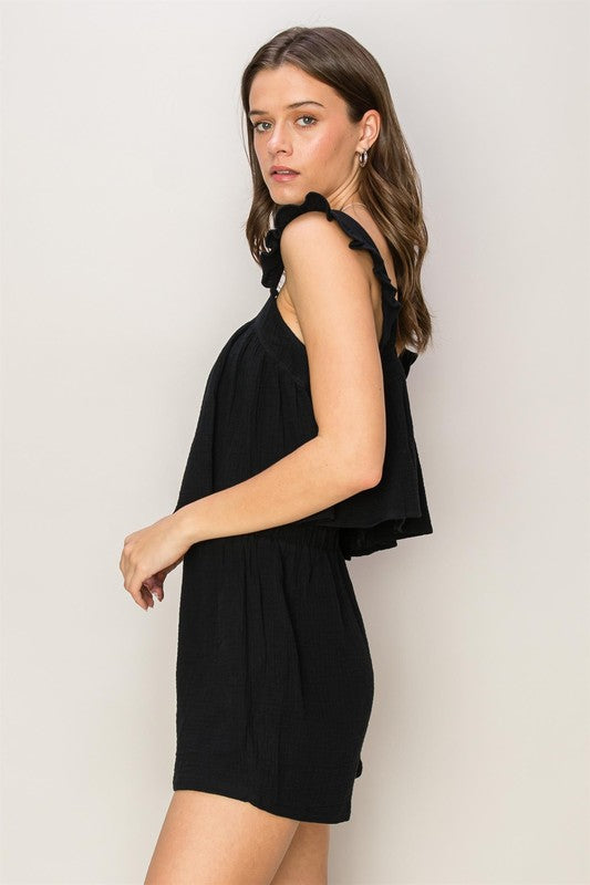 A person stands against a plain background, wearing the Double Gauze Top and Shorts Set, featuring a sleeveless black romper with ruffled straps and a stylish square neck.