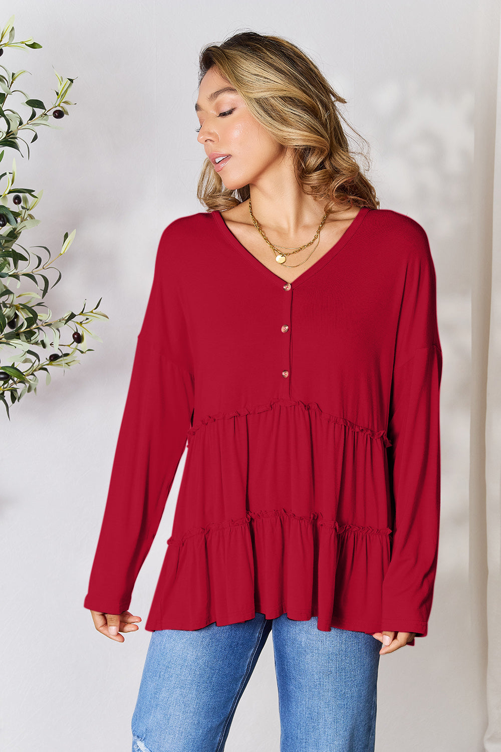 Person stands indoors near a plant, wearing the Double Take Half Button Long Sleeve Ruffle Hem Blouse in gray and blue jeans, smiling while looking to the side. The slightly stretchy fabric of the blouse adds comfort while its buttoned design offers a touch of elegance.