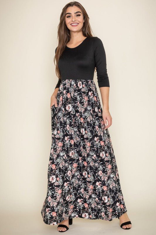 A woman elegantly stands in a room with wooden flooring and modern furniture, wearing a ditsy floral maxi dress and a long-sleeve navy top.