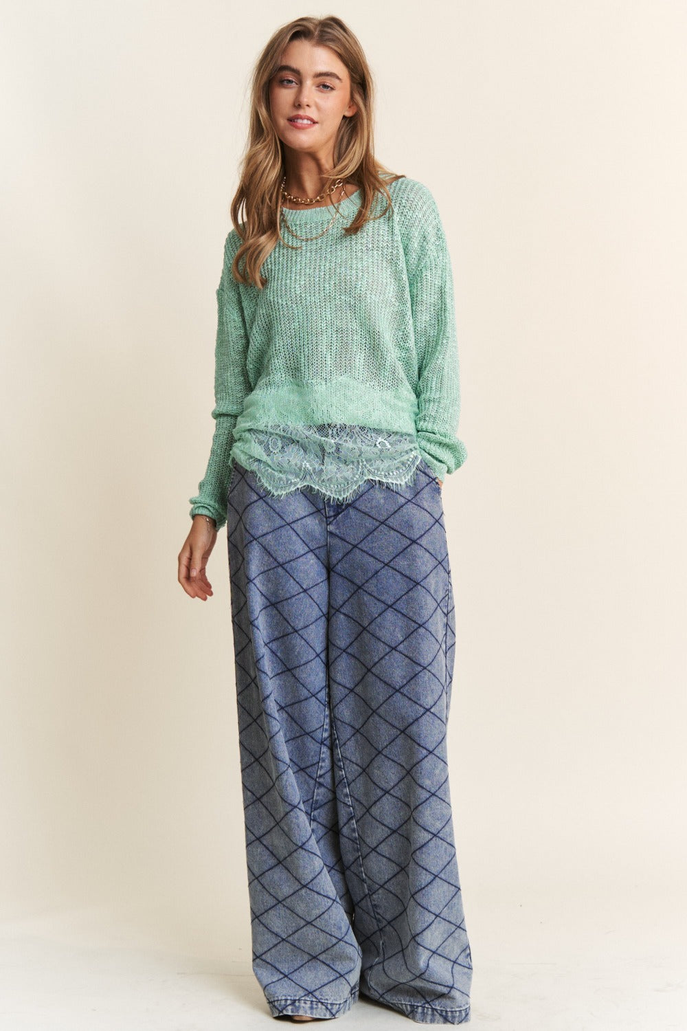 A woman with long hair is wearing the J.NNA Lace Detail Long Sleeve Knit Top in a versatile light green, paired with a lace trim shirt underneath and patterned blue pants. She has a neutral expression and her right hand near her face.