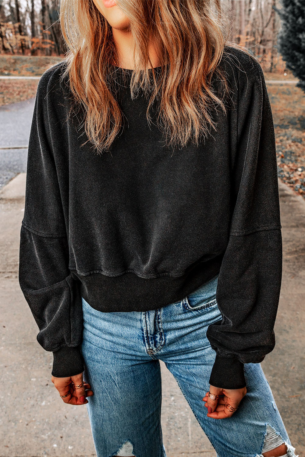 A person with wavy, shoulder-length hair wearing a Black Acid Wash V-shape Open Back Sweatshirt and distressed blue jeans stands outside on a paved surface.