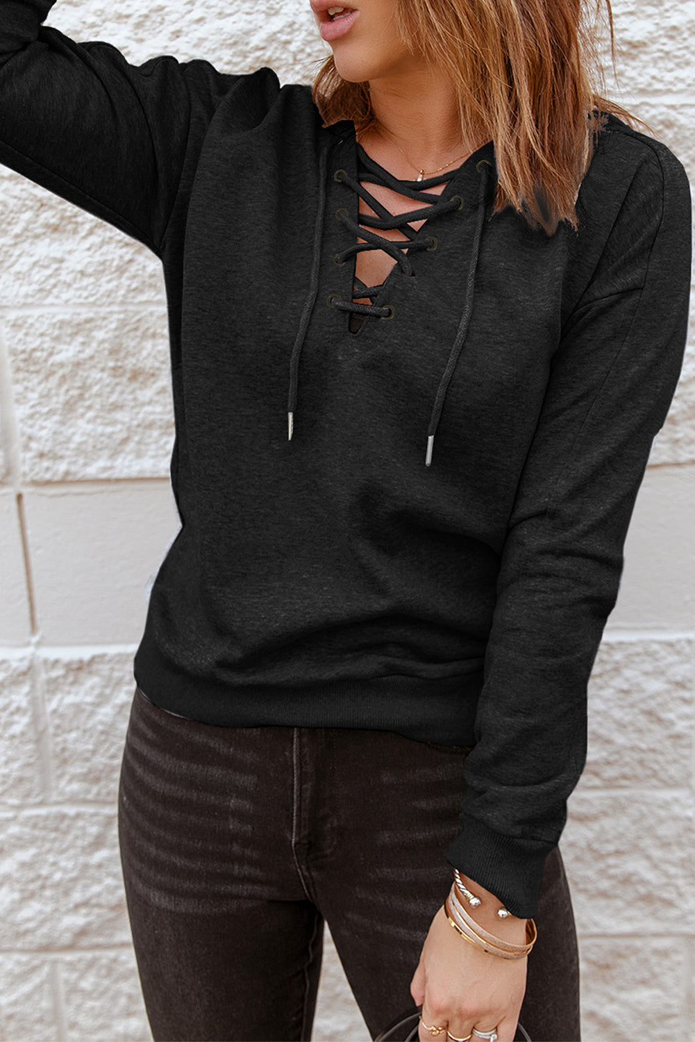 A person with long hair facing away from the camera, wearing a Black Casual Solid Color Lace-up Hoodie and black jeans, standing in front of a white brick wall, epitomizing fashionable comfort.