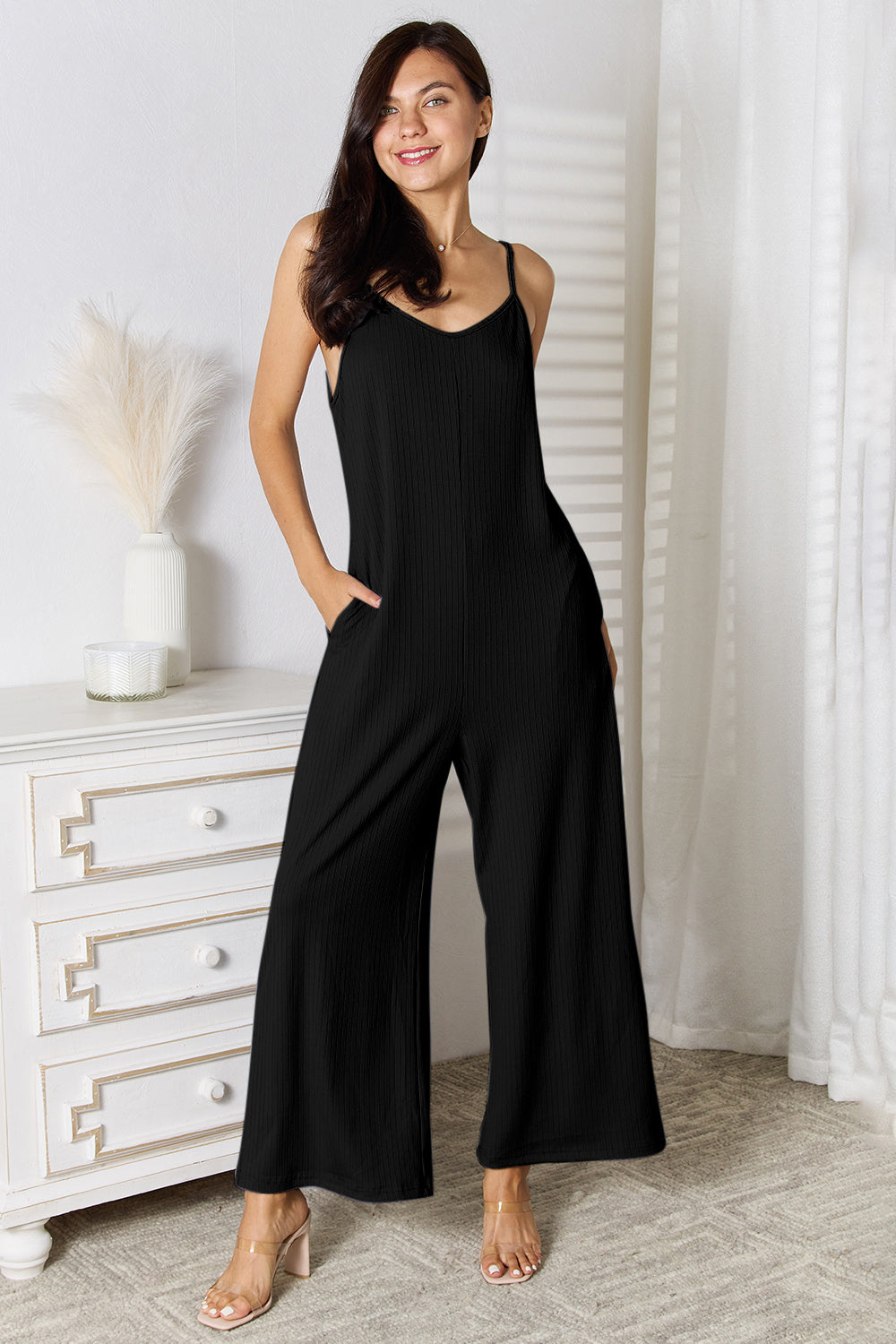 A woman in a stylish Basic Bae Full Size Spaghetti Strap V-Neck Jumpsuit stands indoors near a white dresser adorned with decorative items, smiling with one hand in her pocket.