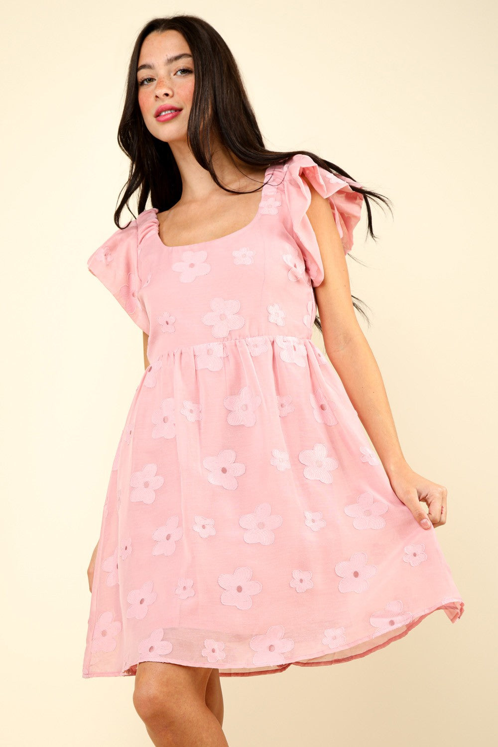 Woman in a VERY J Flower Embroidered Organza Mini Dress with flutter sleeves stands against a light background, resting one hand on the skirt and the other slightly bent. She is wearing strappy sandals.