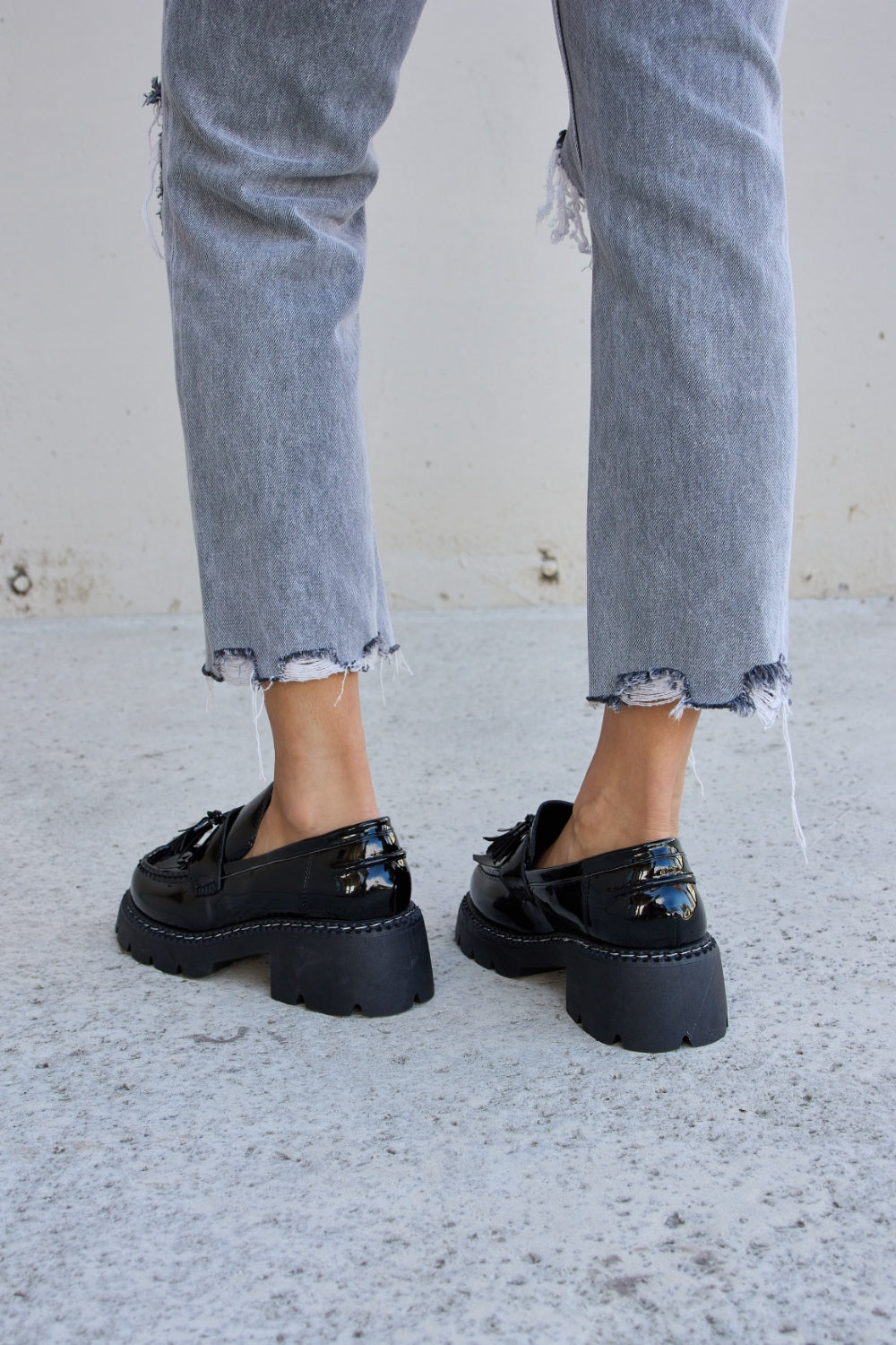 A person wearing ripped gray jeans and the Forever Link Tassel Detail Mid Heel Chunky Loafers, which feature shiny black tones, chunky soles, and a subtle tassel detail.