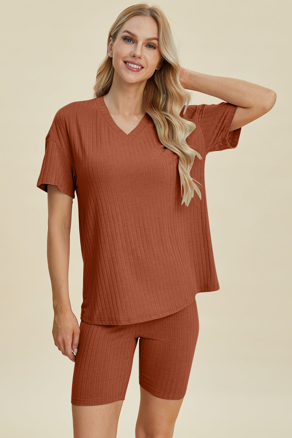 A woman with long blonde hair wearing the Basic Bae Full Size Ribbed V-Neck Short Sleeve Top and Shorts Set in rust stands against a plain background. This stylish two-piece set is made from a comfortable, stretchy fabric that is also machine washable.