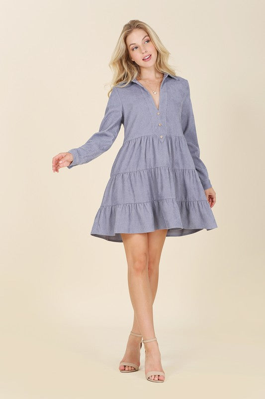 The woman in the Corduroy tiered dress, featuring a deep V-neck, smiles as she gently touches her forehead, showcasing the light blue fabric cascading elegantly.