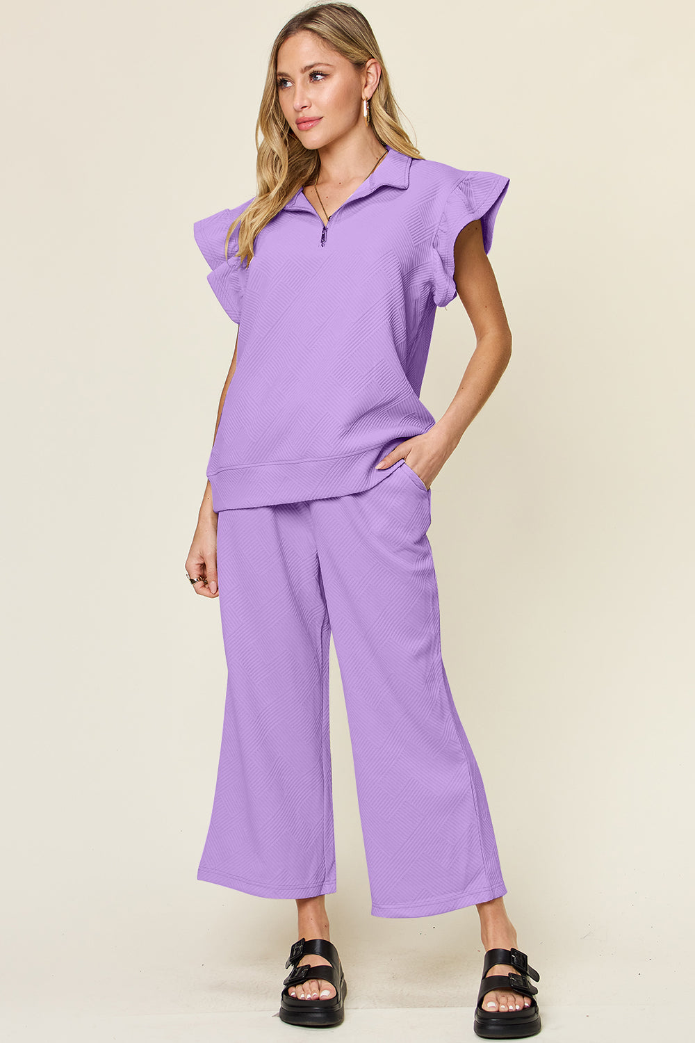 A woman stands wearing the Double Take Texture Ruffle Short Sleeve Top and Drawstring Wide Leg Pants Set in a slightly stretchy, light gray fabric with zipper detail. The two-piece outfit is complemented by black sandals, and she has one hand in her pocket. Machine wash cold for easy care.