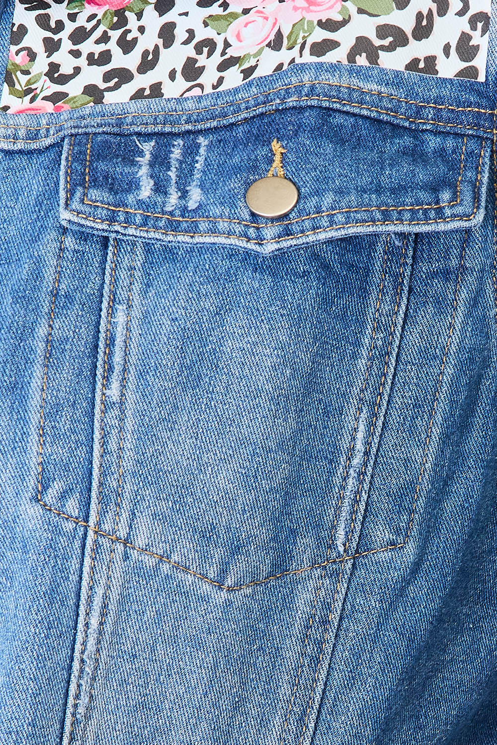 A person smiles while wearing the Baeful Leopard Floral Distressed Raw Hem Denim Jacket with embroidered patches, paired with a white shirt and blue jeans, as they stand indoors near a white piece of furniture.