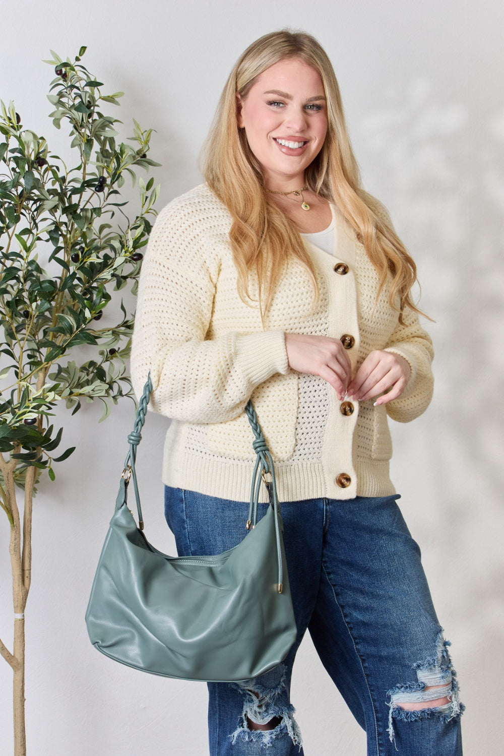 A person in a knitted sweater and jeans holds the SHOMICO Braided Strap Shoulder Bag, a blue-green statement accessory featuring straps crafted from vegan leather.