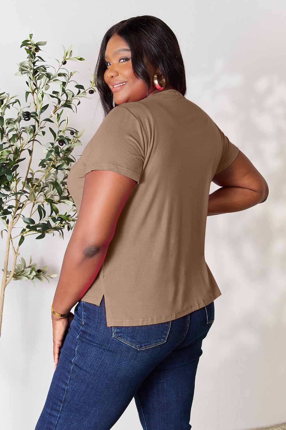 A woman stands next to an indoor plant, smiling, with one hand in her khaki pants pocket. In her pink Basic Bae Full Size Round Neck Short Sleeve T-Shirt and gold jewelry, she radiates casual elegance.