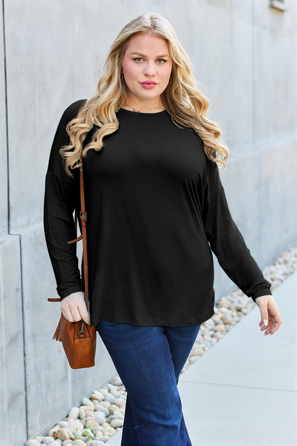 A woman with shoulder-length hair wearing a Basic Bae Full Size Round Neck Dropped Shoulder T-Shirt in green and a pair of jeans, exuding a basic style, stands against a concrete wall. She's holding an imported brown woven backpack on her shoulder while touching her hair with her hand.