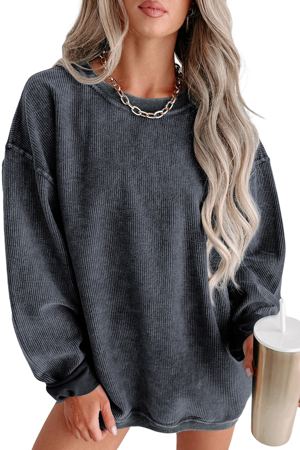 A person with long, blonde hair smiles while wearing an oversized Gray Solid Ribbed Knit Round Neck Pullover Sweatshirt and a gold chain necklace. They are standing indoors near a window, embodying versatile style.