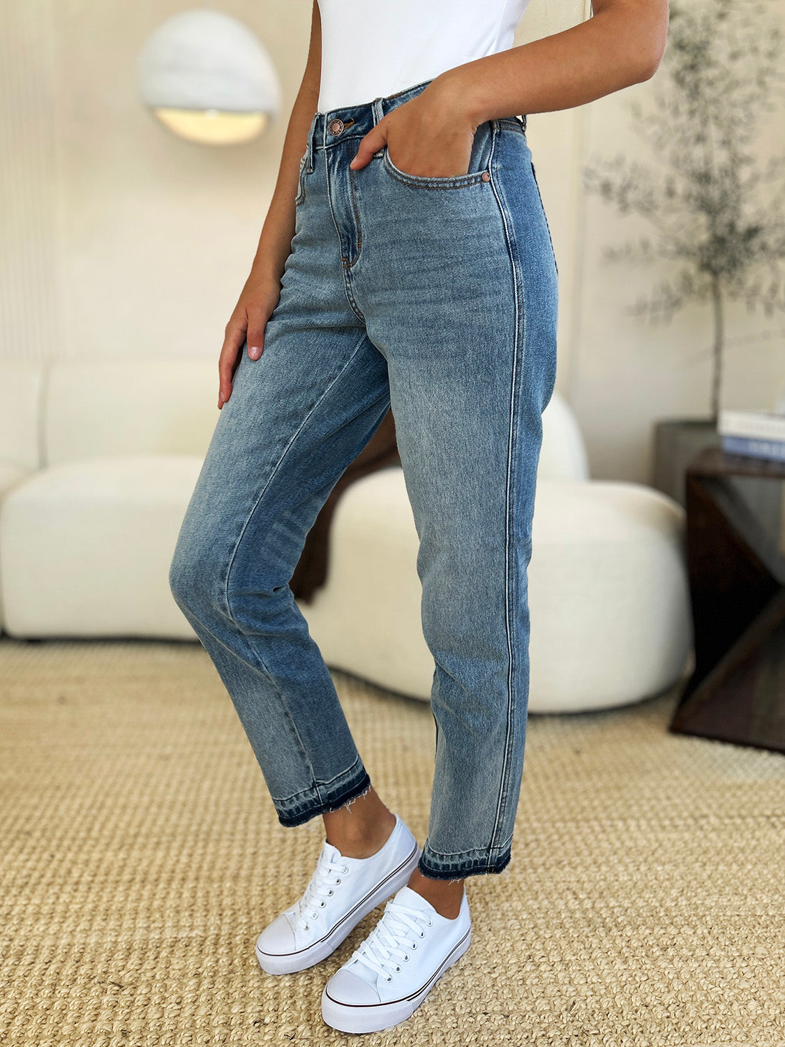 Person wearing a black top, Judy Blue Full Size Mid Rise Rigid Magic Release Hem Jeans, and black sneakers with white laces, standing on a beige carpet with hands in pockets.