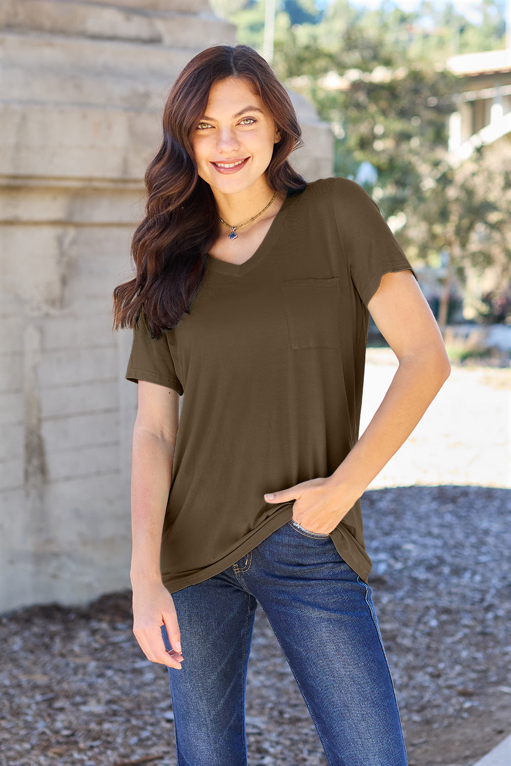 A woman with long brown hair, wearing a Basic Bae Full Size V-Neck Short Sleeve T-Shirt in teal and stretchy blue jeans, stands outdoors near a stone structure, smiling at the camera with one hand in her pocket.