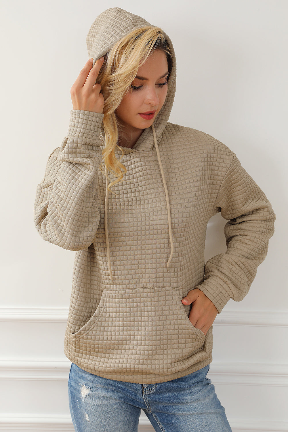 Rear view of a person with blonde hair wearing a Khaki Quilted Kangaroo Pocket Drawstring Hoodie and blue jeans, standing against a white wall.