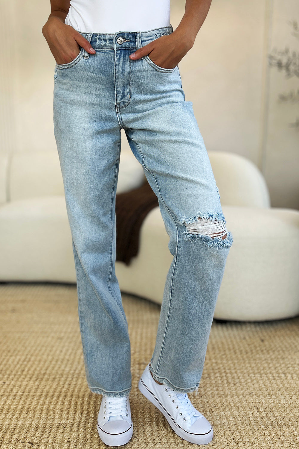 A person standing on a beige carpet, wearing Judy Blue Full Size High Waist Distressed Straight Jeans in light blue, a white top, and white sneakers.