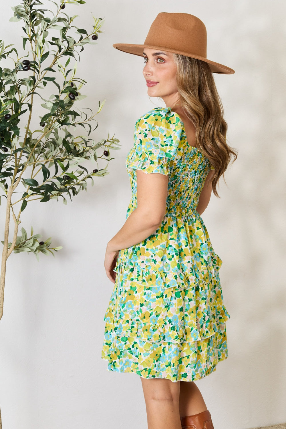 A woman wearing the Devine Smocked Scoop Neck Flounce Sleeve Mini Dress and a brown hat smiles and poses near greenery indoors.