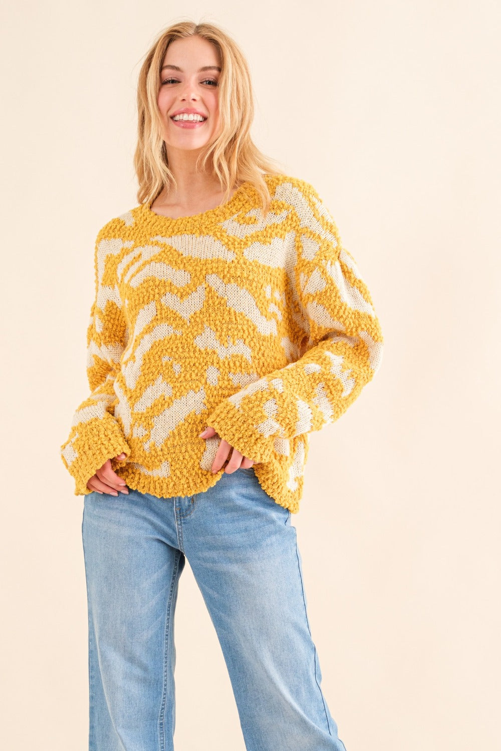 A person with long hair wearing a stylish yellow and white And The Why Full Size Textured Pattern Contrast Sweater and blue jeans stands against a beige background.