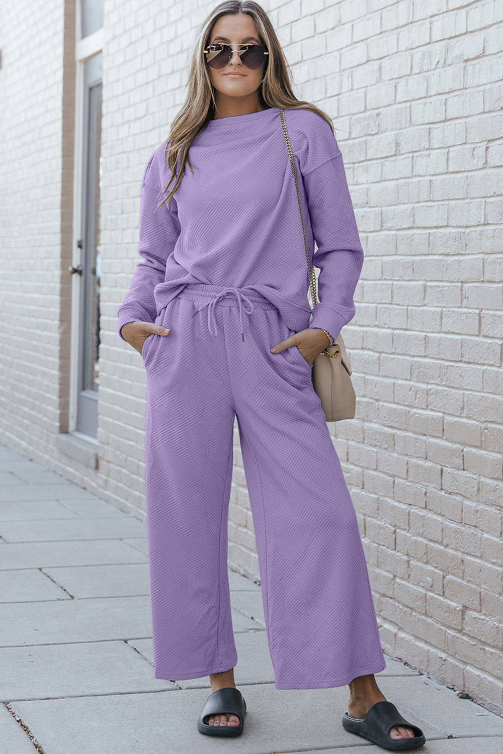 A woman with blonde hair, dressed in the Double Take Full Size Textured Long Sleeve Top and Drawstring Pants Set featuring a stretchy black long-sleeve top and matching wide-leg trousers made of rayon spandex, stands barefoot in a room with white walls and a wooden floor.