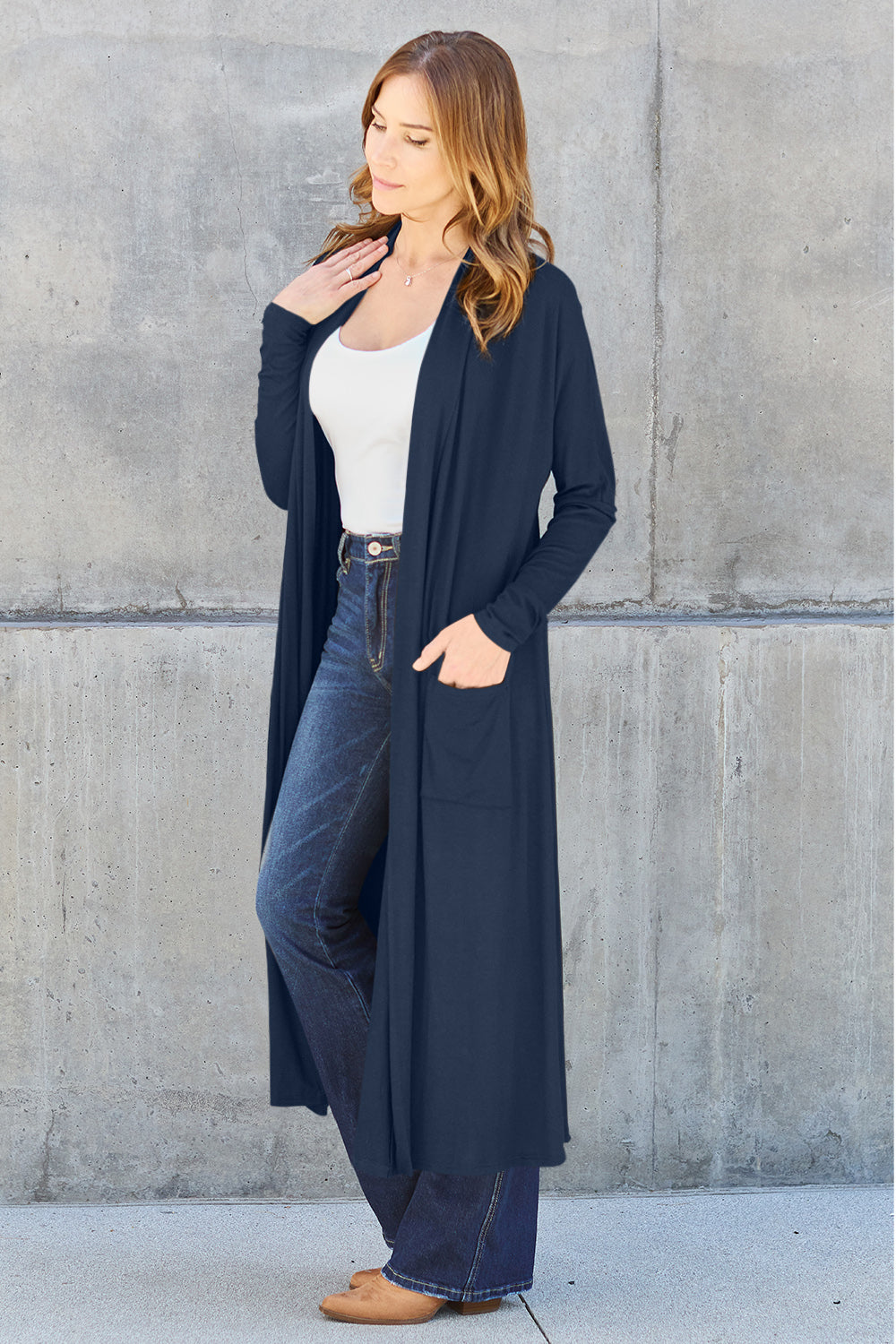 A woman stands against a concrete wall, wearing the Basic Bae Full Size Open Front Long Sleeve Cover Up made from rayon spandex, a white top, blue jeans in her perfect size, and brown shoes with her hands in her pockets. Machine wash cold for best results.