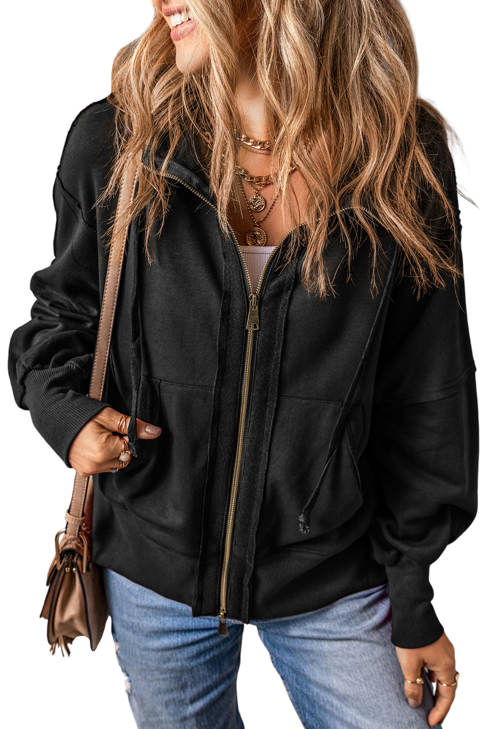 A person with long, wavy blonde hair is shown from the back, wearing a Black Raw Edge Exposed Seam Full Zip Hoodie and blue jeans, and carrying a beige shoulder bag.