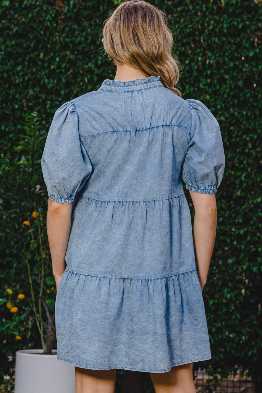 A person wearing the ODDI Washed Tiered Mini Denim Dress with puffed sleeves stands in front of a green leafy background.