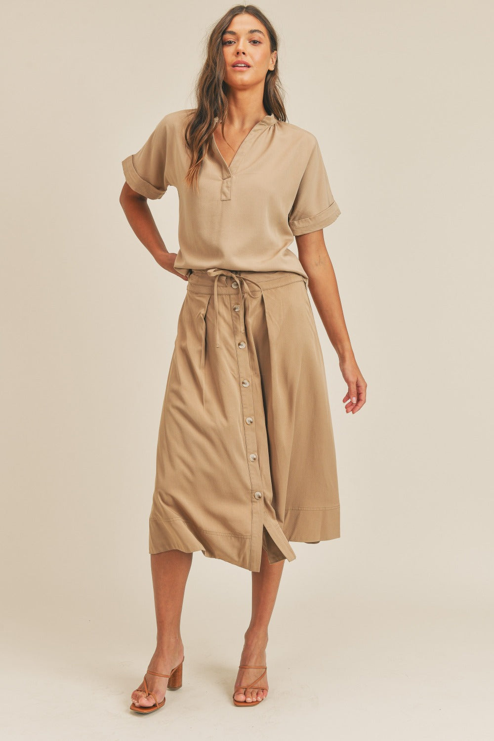 A woman is standing against a plain background wearing the MABLE Short Sleeve Top and Button Down Midi Skirt Set in beige. The elegant two-piece set, which includes a belted midi skirt, is paired perfectly with beige heels as she confidently poses with one hand on her hip.