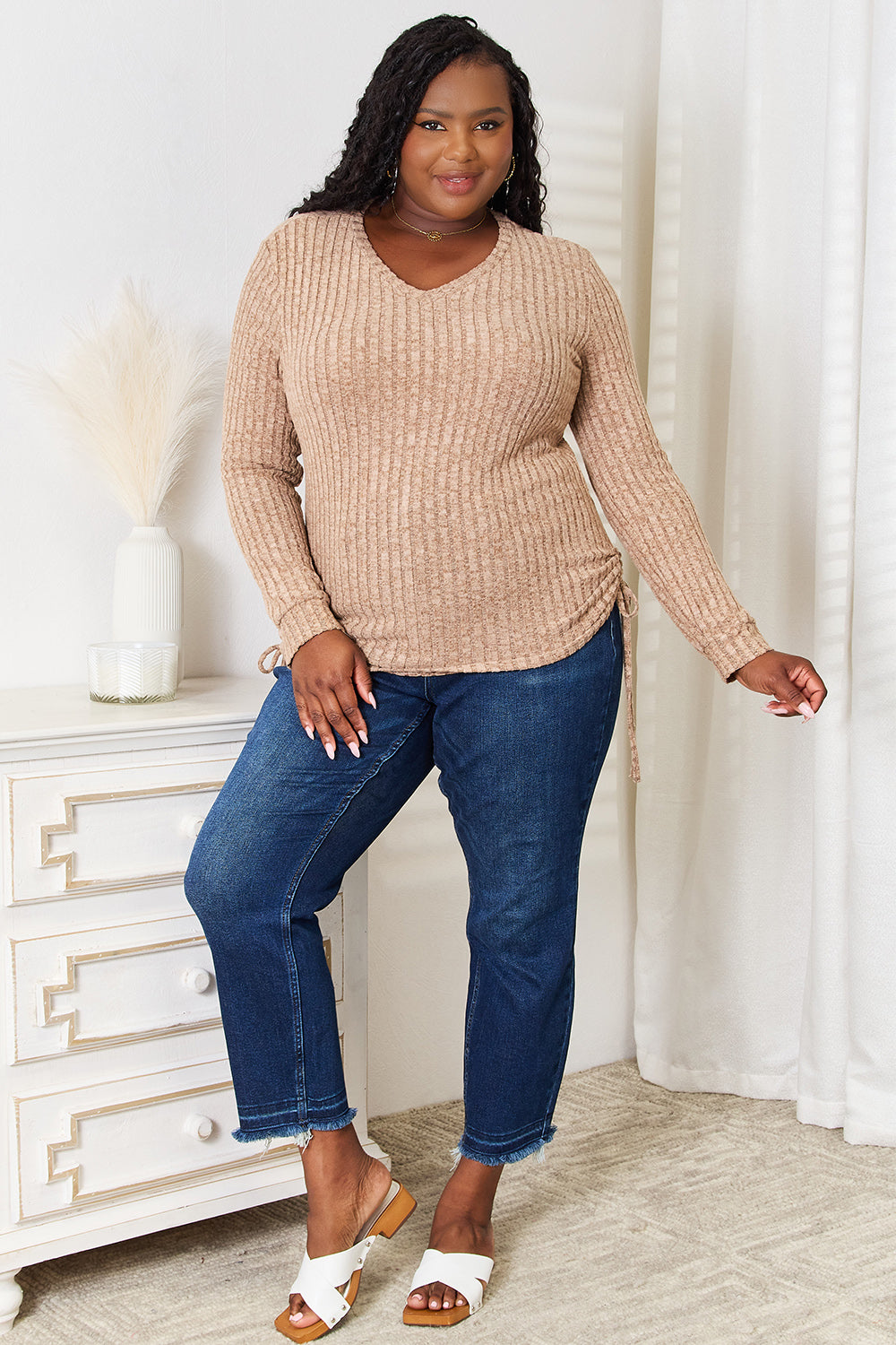 A woman stands indoors against a neutral background, wearing the "Double Take Drawstring Ribbed Long Sleeve T-Shirt" in light purple paired with dark pants. She is smiling and posing with one hand on her hip.