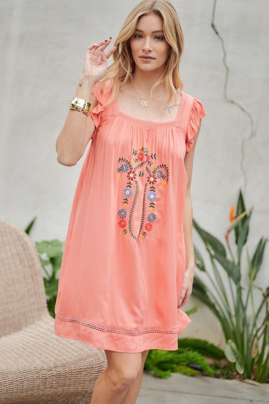 A woman in an Embroidered Square Neck Mini Dress, featuring a coral hue, stands near plants adjusting her hair—the perfect summer attire.