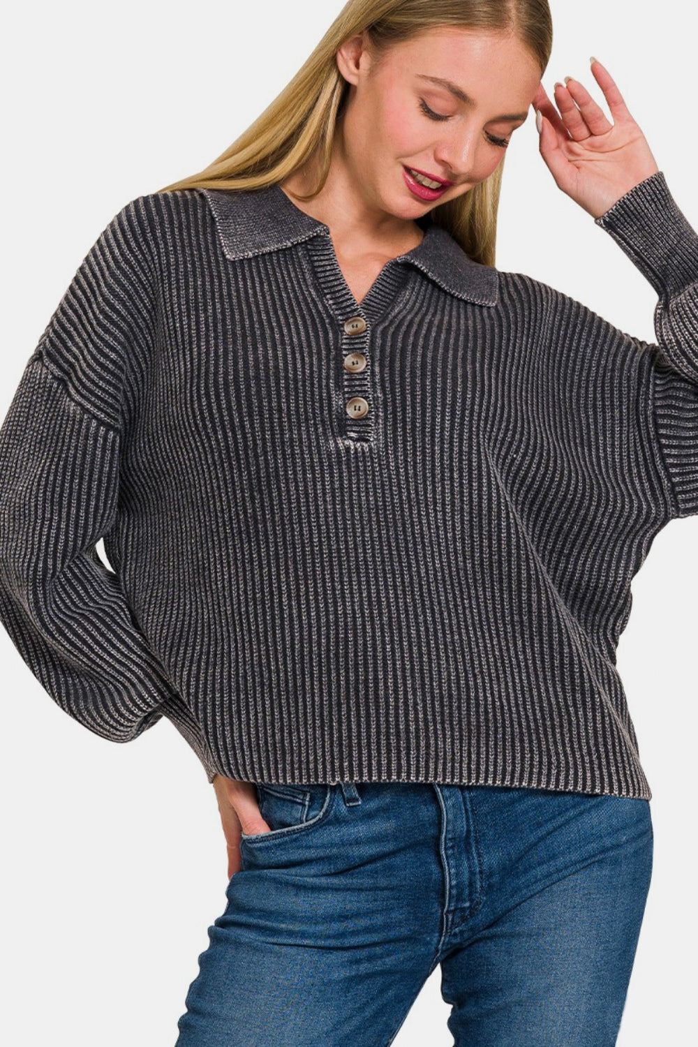 A woman with long blonde hair modeling the Zenana Washed Half Button Long Sleeve Sweater, made of soft washed fabric in grey, paired with blue jeans against a plain backdrop.