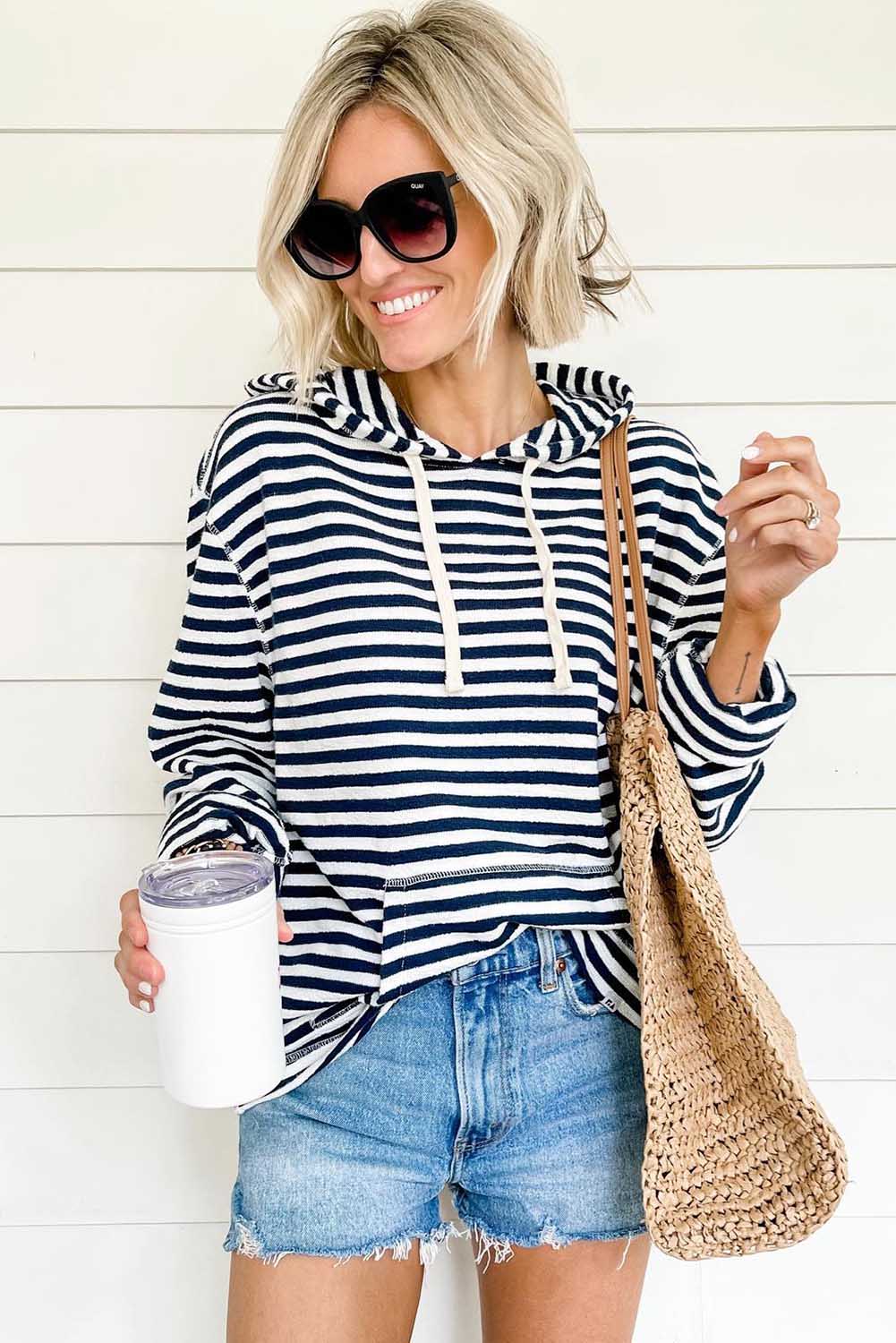 A person with long, blonde hair wearing a trendy Stripe Kangaroo Pocket Drawstring Hoodie with black and white stripes and dark green pants is standing outdoors, facing away from the camera.