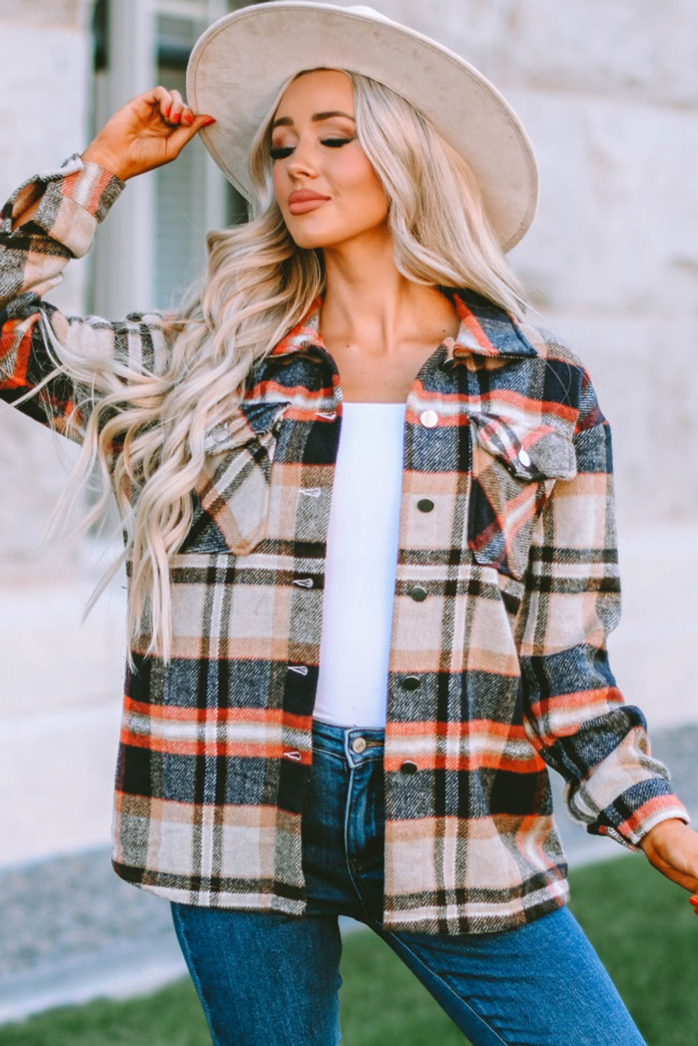 A person wearing a Brown Plaid Button Up Long Sleeve Flannel Shacket layered over a white top with ripped jeans and holding a brown handbag stands against a textured gray wall.