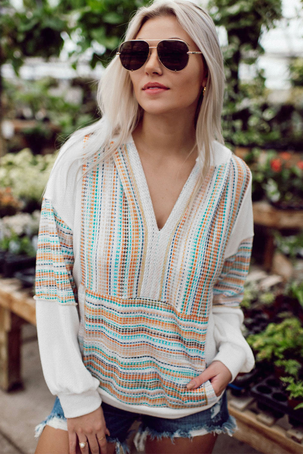 A woman with long blonde hair wearing a Multicolor Striped Patchwork V Neck Drop Shoulder Knit Hoodie and denim shorts stands in a plant nursery. She has sunglasses perched on her head and is looking down.