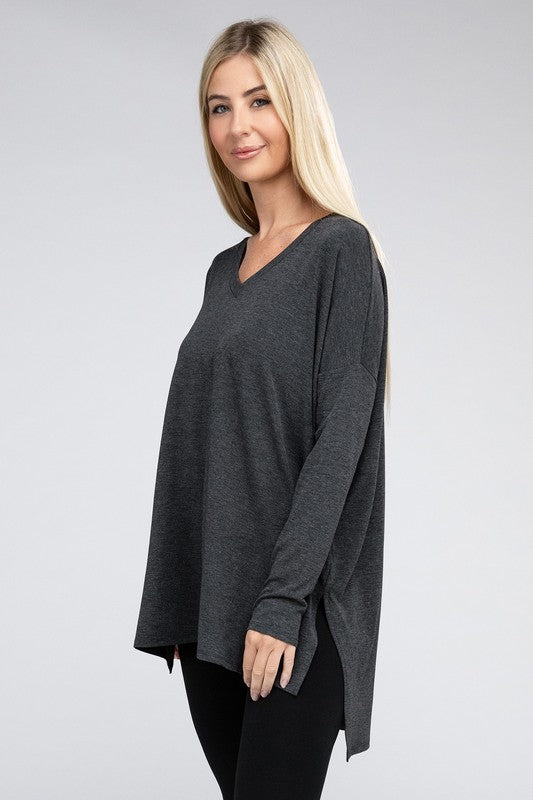 A woman with long, blonde hair is wearing a loose-fitting, brown Dolman Long Sleeve V-Neck Side Slit Hi-Low Hem Top and black pants. She poses with one hand on her hip and a neutral facial expression against a light grey background.