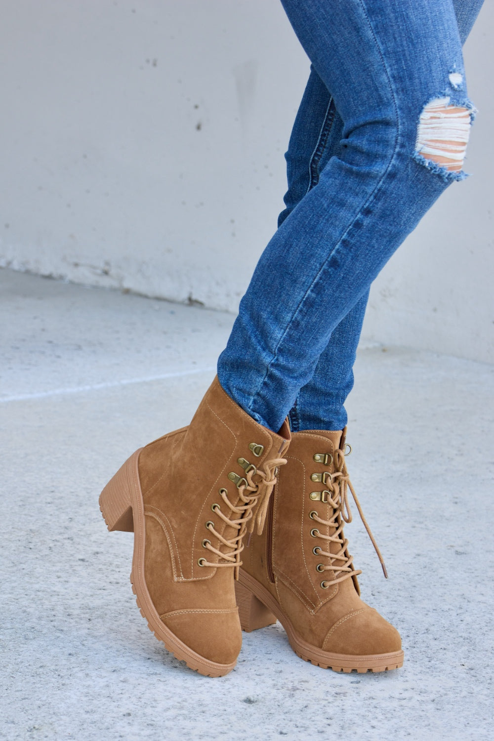 A person wearing distressed blue jeans and Forever Link Lace-Up Zipper Detail Block Heel Boots stands on a concrete surface.