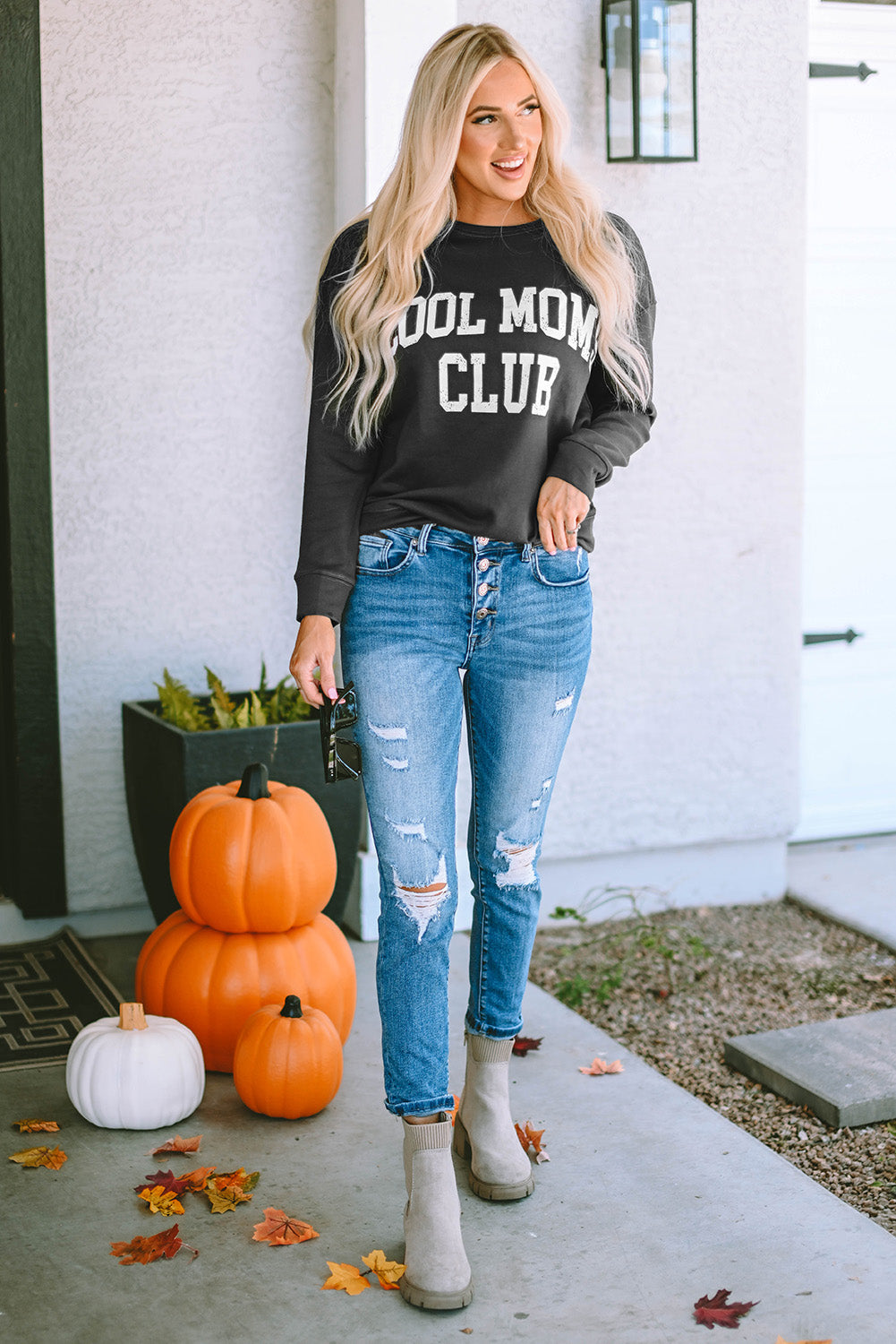 A person with long blond hair wearing a gray COOL MOMS CLUB drop shoulder sweatshirt and relaxed-fit blue jeans is seen from the back, standing near a pumpkin and a window.