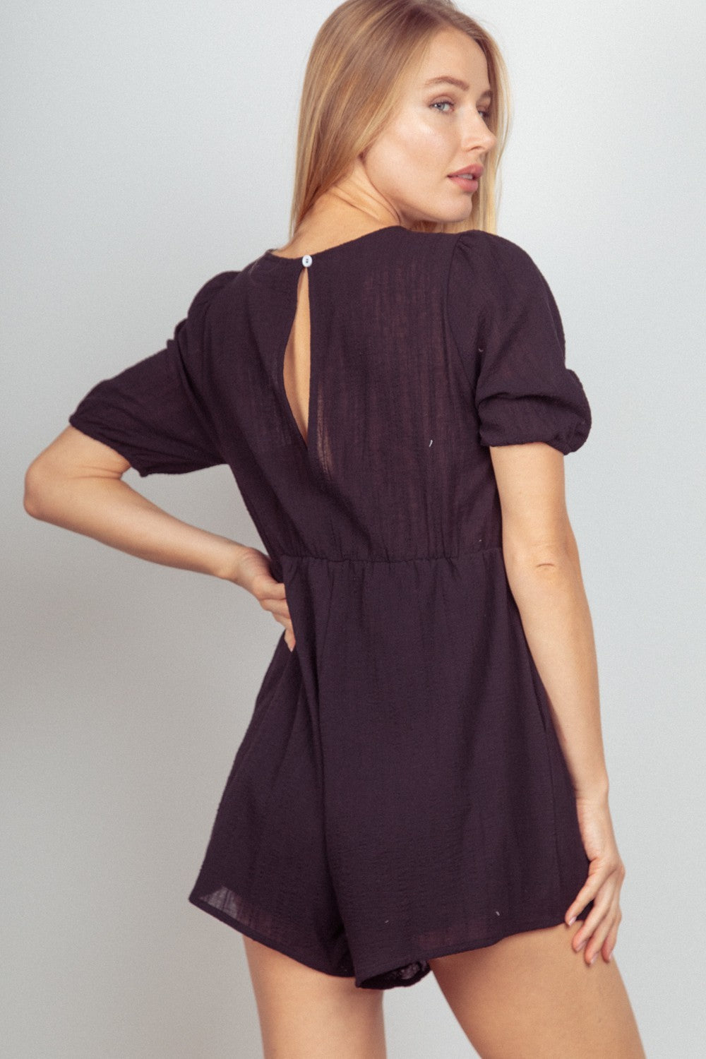 A woman with long blond hair stands against a neutral background, smiling with her hands in the pockets of her black VERY J Lace Detail Puff Sleeve Romper.