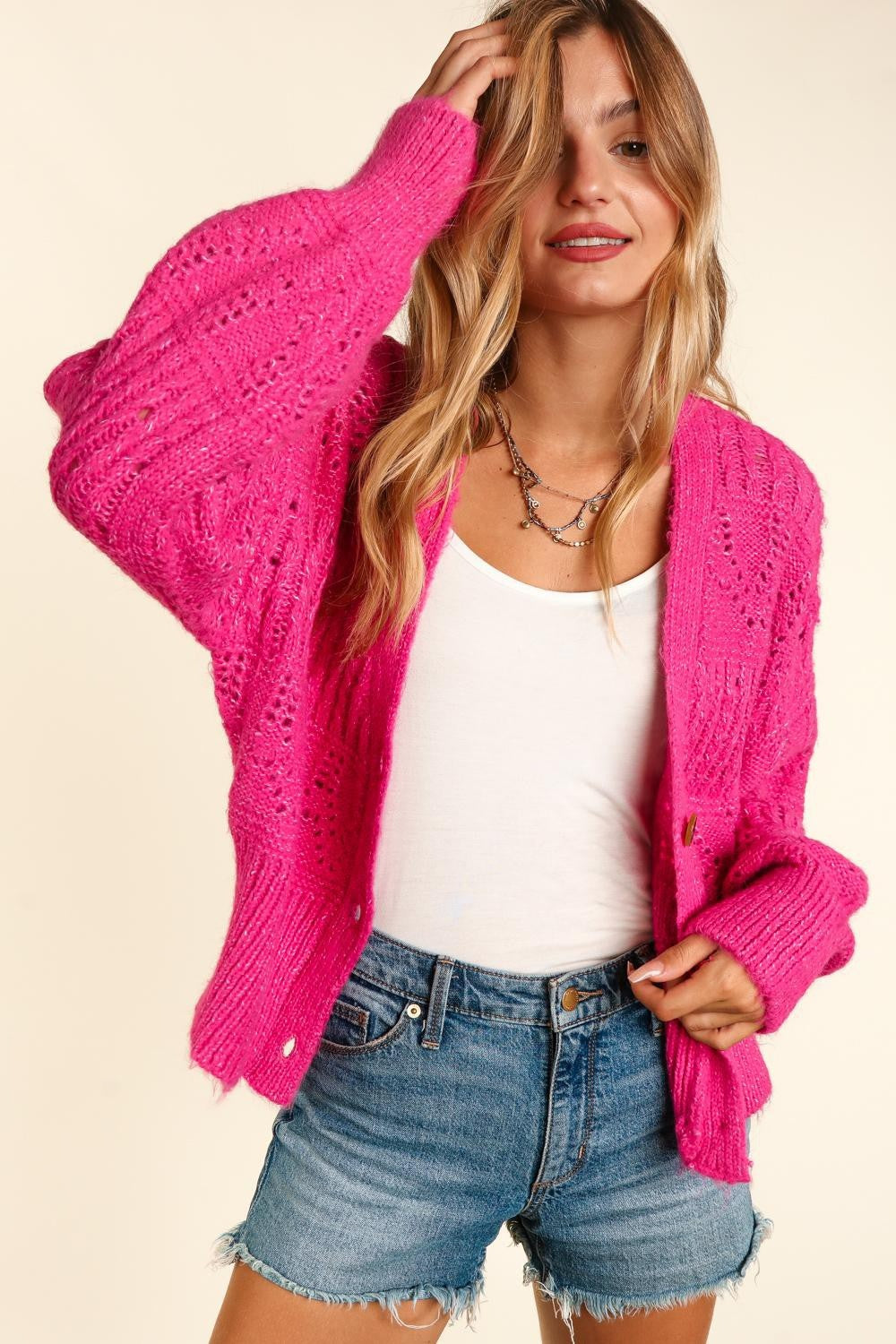 A person with long hair wears the trendy, bright pink Haptics Full Size Button Down Crop Long Sleeve Cardigan over a white top and denim shorts. They hold one side of the cardigan's collar and smile at the camera, showcasing their stylish and versatile layering.